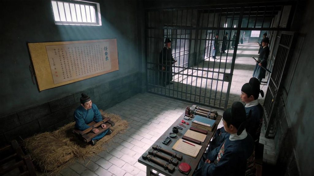 •Lan Yu in chains inside a Ming Dynasty imperial prison, being interrogated by officials, with the “Record of Treasonous Officials” scroll on the wall, in a grim and oppressive atmosphere.
